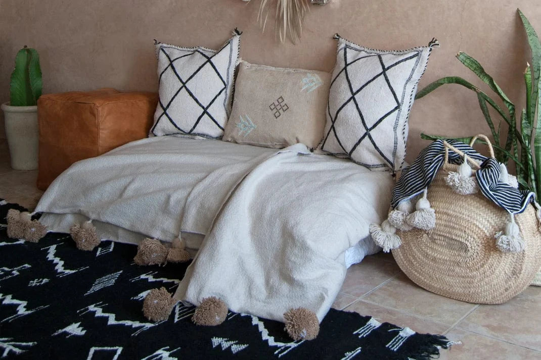 Decorative pillows and blanket on a bed with a woven basket and plants in the background.